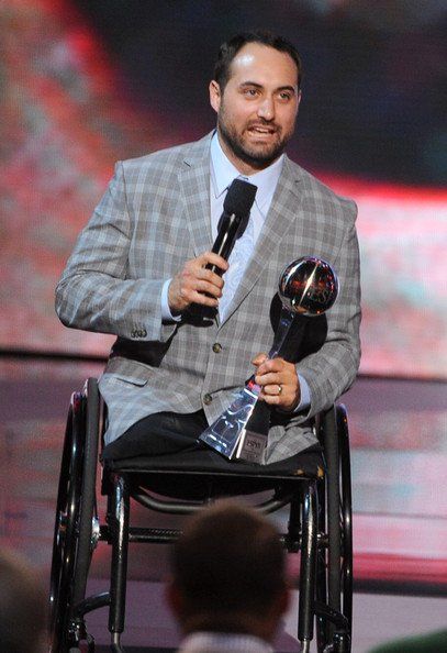 Josh Sweeney on stage in his wheelchair, holding a microphone and an award.